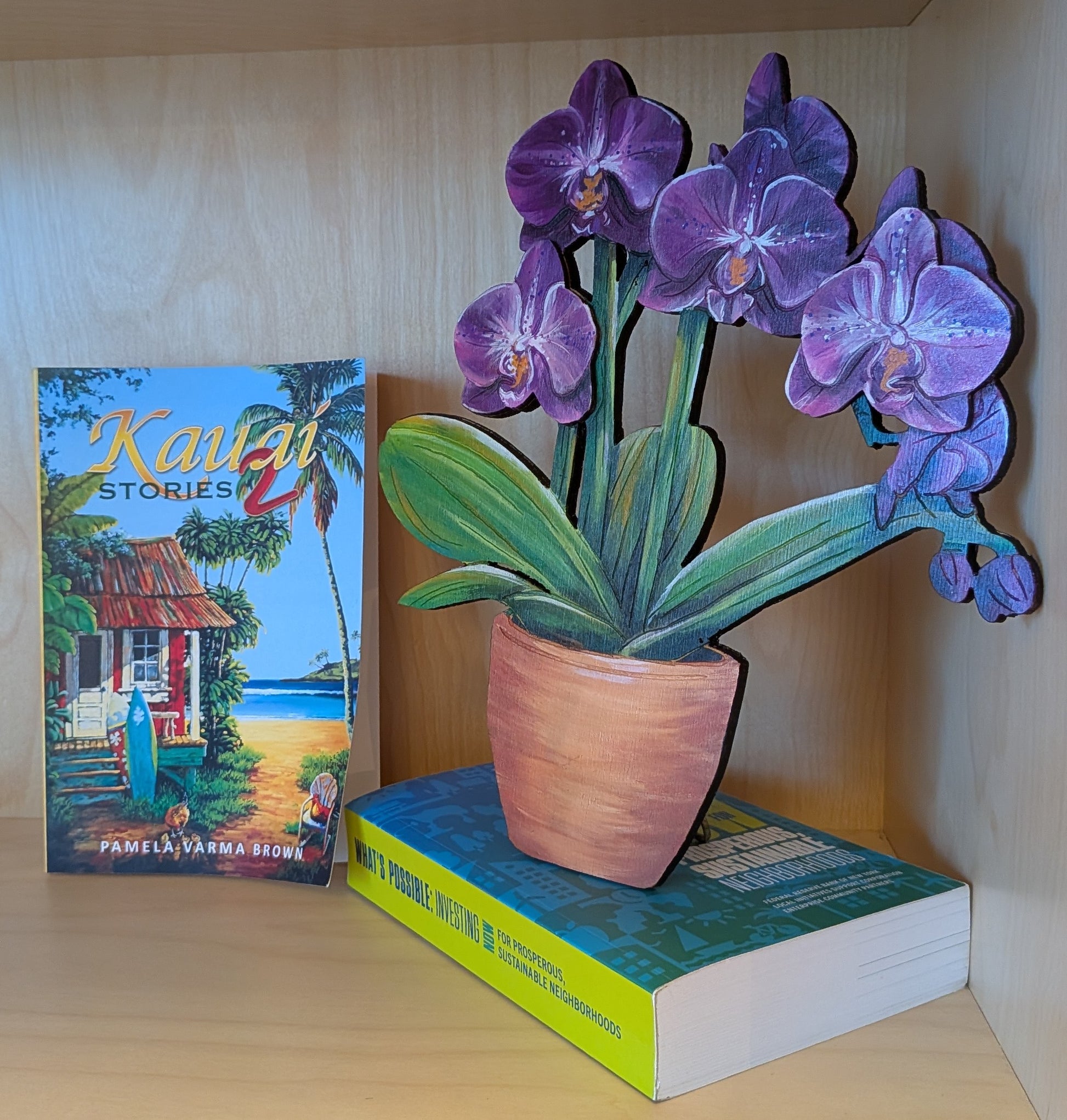 A decorative wooden cutout of purple orchids in a terracotta pot displayed on top of a book on a wooden shelf, next to a book titled 'Kailua Stories'.